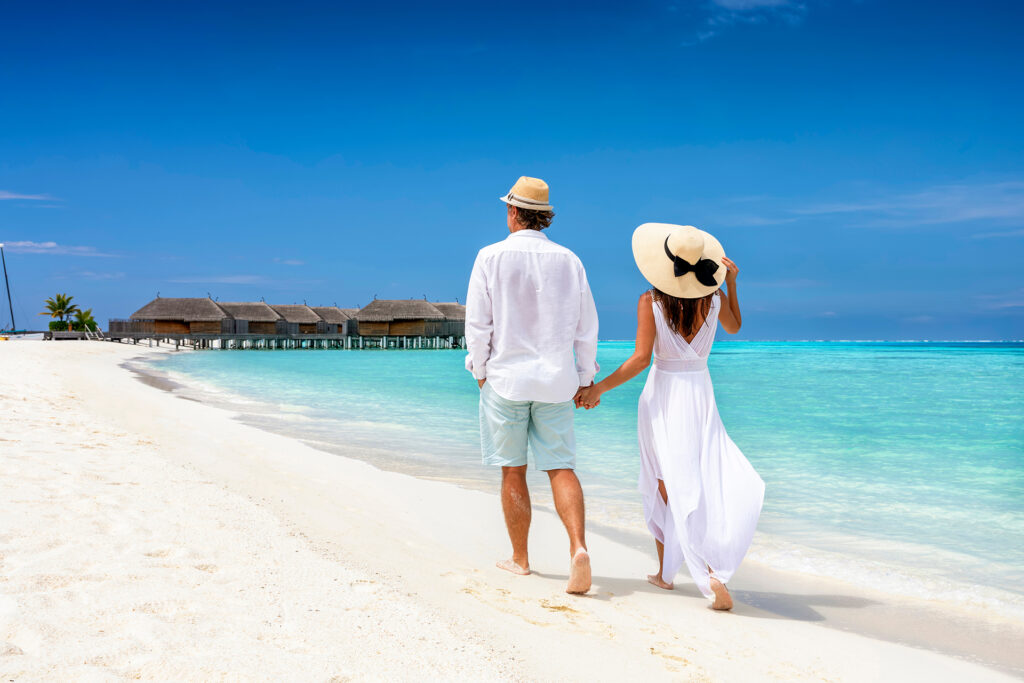 A happy couple wearing white clothing and hats walking along a beautiful tropical beach with turquoise water.